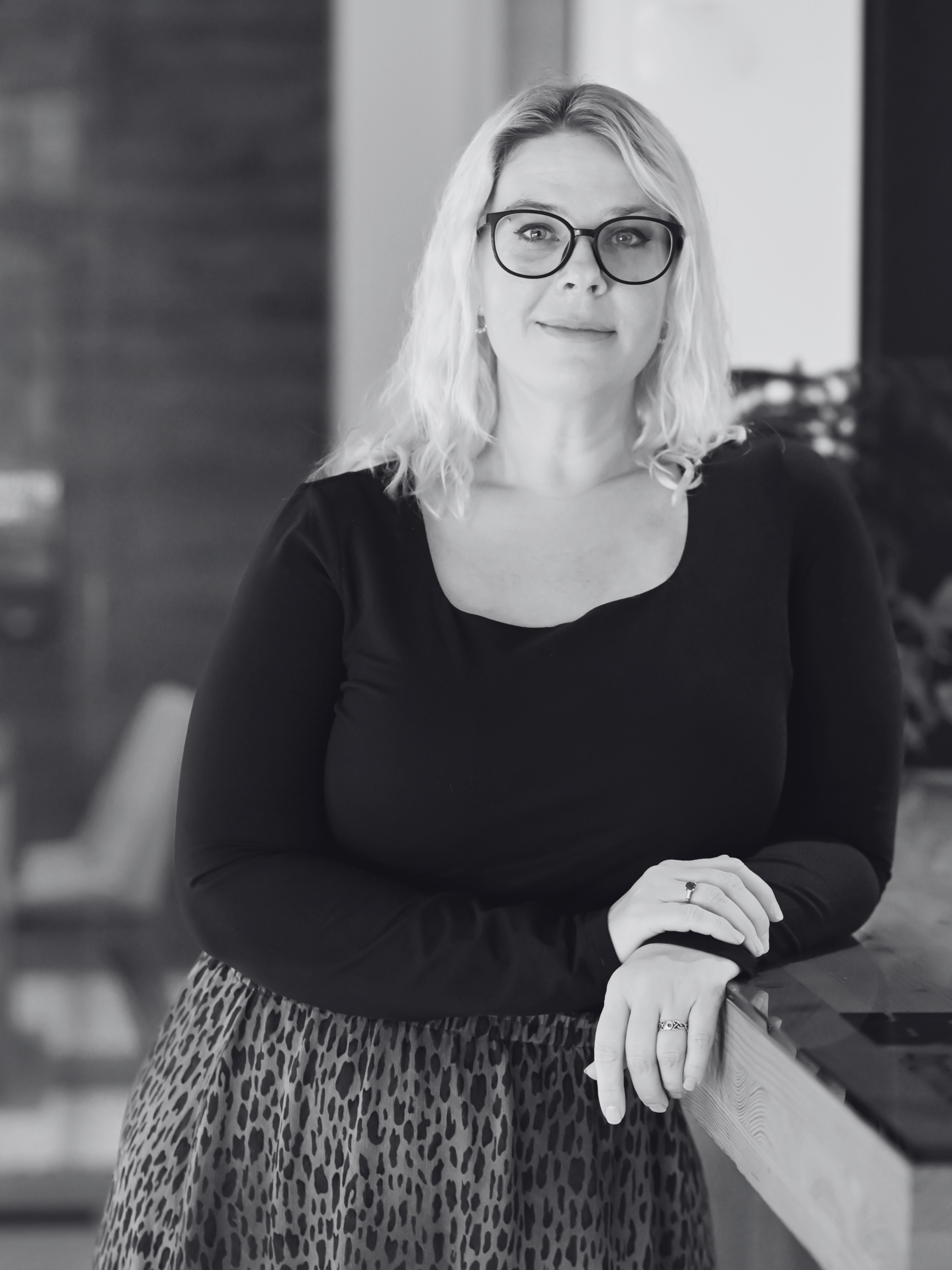Black and white photograph of a woman with blonde hair, dark rimmed glasses and a dark top leaning professionally on a counter